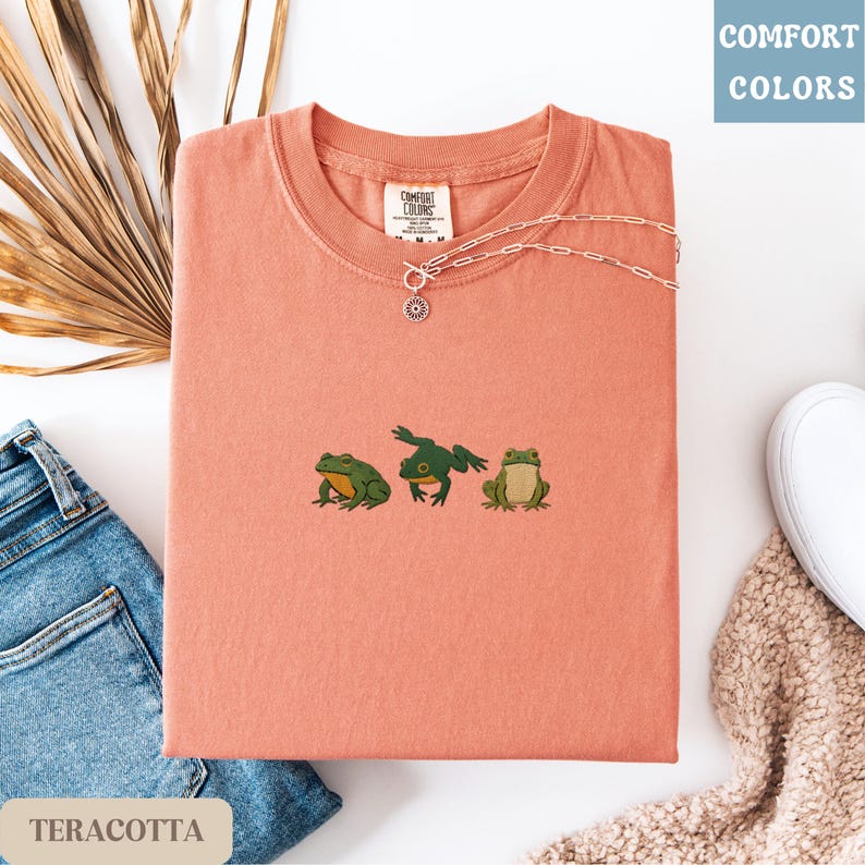 May include: Terracotta-colored t-shirt with three embroidered frog designs. A silver necklace with a sun pendant rests on the shirt. The shirt is folded with a pair of jeans and a white shoe in the background. The text "COMFORT COLORS" is in the upper right corner.