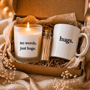May include: A gift box containing a lit candle in a glass jar with the text no words. just hugs., a small glass vial filled with wooden matches, and a white ceramic mug with the word hugs. printed in black on the side, nestled in paper.