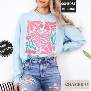 May include: Light blue long-sleeve shirt with a floral and heart graphic. The design includes pink flowers, a zebra-striped heart, cherries, and the text "Comfort Colors" and "Chambray". The shirt is made of a soft material.
