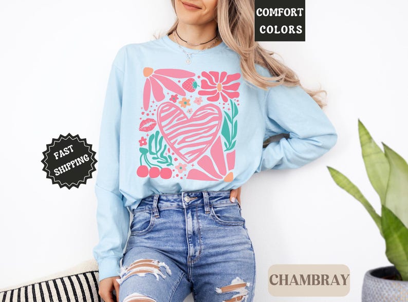 May include: Light blue long-sleeve shirt with a floral and heart graphic. The design includes pink flowers, a zebra-striped heart, cherries, and the text "Comfort Colors" and "Chambray". The shirt is made of a soft material.