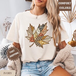 May include: Ivory-colored t-shirt featuring a large, patchwork-style leaf design in shades of brown, orange, and cream. The leaf design has various patterns. The words "COMFORT COLORS" are printed on the top right. The word "IVORY" is printed on the bottom left.