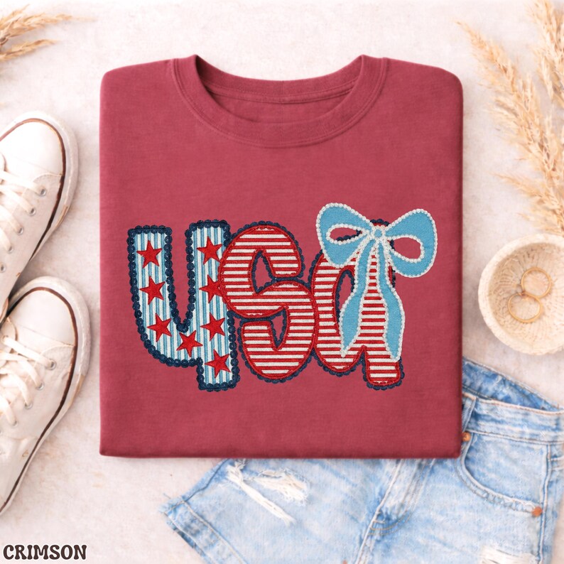 Op de afbeelding: Karmizijnrood T-shirt met het woord "USA" in een patriottisch ontwerp. De "U" is blauw met rode sterren, de "S" en "A" zijn rood en wit gestreept en een blauwe strik staat rechts. Witte sneakers en een spijkerbroekshort op de achtergrond.