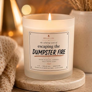 Op de afbeelding: Een brandende kaars in een matglazen pot met een etiket waarop staat: SMELLS LIKE the calming scent of escaping the DUMPSTER FIRE. Het etiket bevat tonen van verlichting, vernieuwing, 312g, 100% sojawas, met de hand gegoten.
