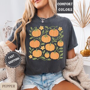 May include: Dark gray t-shirt featuring a pumpkin graphic with orange pumpkins, green vines, and yellow flowers. The shirt has the words "COMFORT COLORS" printed on it. The shirt is made of a soft material.