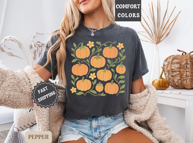 May include: Dark gray t-shirt featuring a pumpkin graphic with orange pumpkins, green vines, and yellow flowers. The shirt has the words "COMFORT COLORS" printed on it. The shirt is made of a soft material.