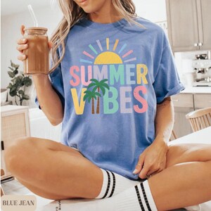 K&ouml;nnte beinhalten: Ein blaues T-Shirt mit den Worten "SUMMER VIBES" in bunten Buchstaben, einer Sonne und Palmen. Das Shirt hat eine lockere Passform und hochgekrempelte &Auml;rmel. Die Person tr&auml;gt wei&szlig;e Socken mit schwarzen Streifen.