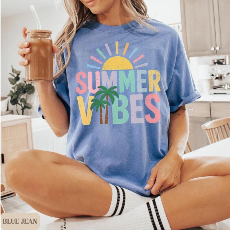 K&ouml;nnte beinhalten: Ein blaues T-Shirt mit den Worten "SUMMER VIBES" in bunten Buchstaben, einer Sonne und Palmen. Das Shirt hat eine lockere Passform und hochgekrempelte &Auml;rmel. Die Person tr&auml;gt wei&szlig;e Socken mit schwarzen Streifen.