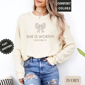May include: Ivory long-sleeve sweatshirt with the text "SHE IS WORTHY" and a bow graphic. The sweatshirt is paired with blue jeans. The words "COMFORT COLORS" and "IVORY" are also visible.