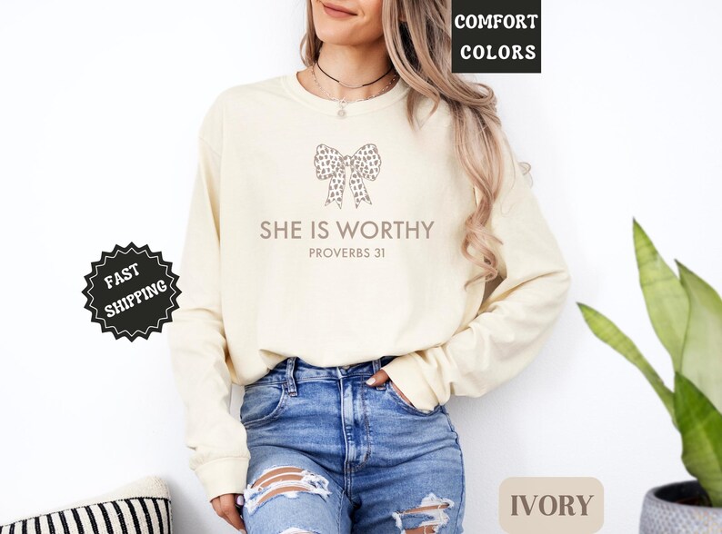 May include: Ivory long-sleeve sweatshirt with the text "SHE IS WORTHY" and a bow graphic. The sweatshirt is paired with blue jeans. The words "COMFORT COLORS" and "IVORY" are also visible.