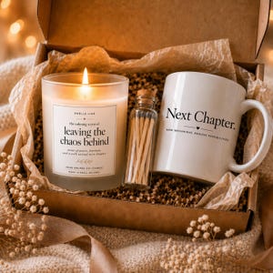 May include: A gift box containing a lit soy candle in a glass jar with a label reading 'Smells like the calming scent of leaving the chaos behind', a small glass bottle filled with wooden matches, and a white ceramic mug printed with 'Next Chapter'.