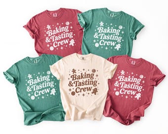 Peut inclure: Cinq t-shirts à manches courtes de couleurs rouge, vert et crème. Chaque t-shirt affiche le texte "Baking & Tasting Crew" dans une couleur contrastante, avec des bonhommes en pain d'épices et des graphismes festifs.