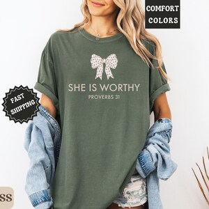 May include: Moss green t-shirt with the text "SHE IS WORTHY" and "PROVERBS 31" in a light color. The shirt features a bow design. The words "COMFORT COLORS" and "FAST SHIPPING" are also visible.