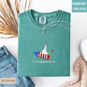 K&ouml;nnte beinhalten: Ein meergr&uuml;nes T-Shirt mit dem gestickten Text "firequacker" unter einer Cartoon-Gans, die auf einer Rakete reitet. Die Gans tr&auml;gt einen patriotischen Hut. Eine silberne Halskette mit Anh&auml;nger liegt auf dem Shirt. Das Shirt ist gefaltet.