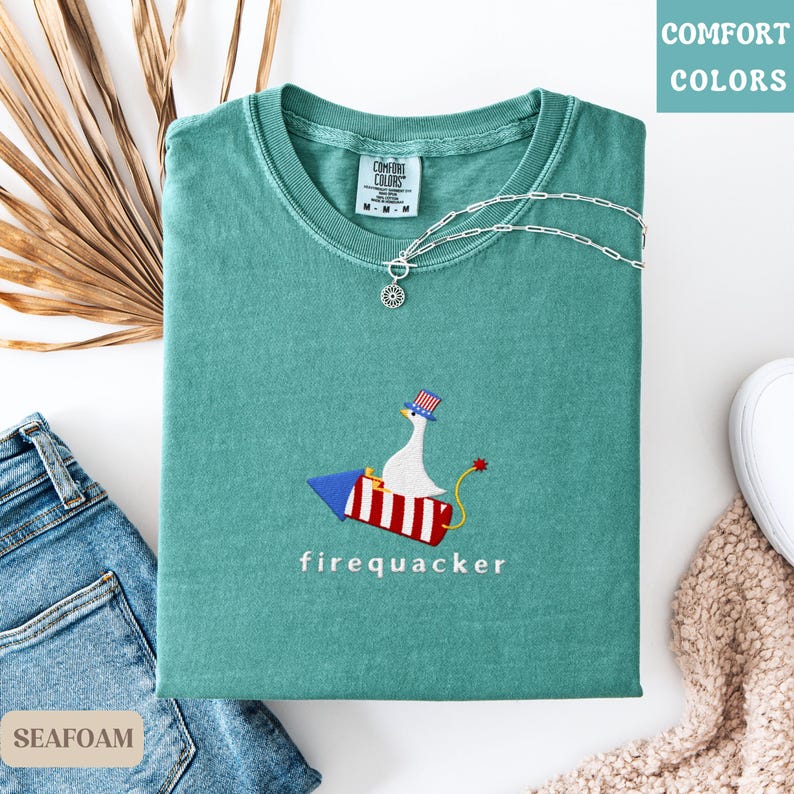 K&ouml;nnte beinhalten: Ein meergr&uuml;nes T-Shirt mit dem gestickten Text "firequacker" unter einer Cartoon-Gans, die auf einer Rakete reitet. Die Gans tr&auml;gt einen patriotischen Hut. Eine silberne Halskette mit Anh&auml;nger liegt auf dem Shirt. Das Shirt ist gefaltet.