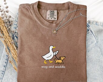 May include: Espresso-colored Comfort Colors t-shirt with a graphic of a duck walking a dog on a leash. The text "wag and waddle" is below the graphic. A silver necklace with a small pendant is on the shirt.