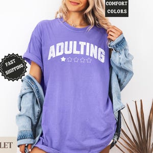 May include: A violet-colored t-shirt with the word "ADULTING" in white block letters. The shirt has five white stars below the word. The model is wearing a denim jacket over the shirt. The words "COMFORT COLORS" are in a black box.