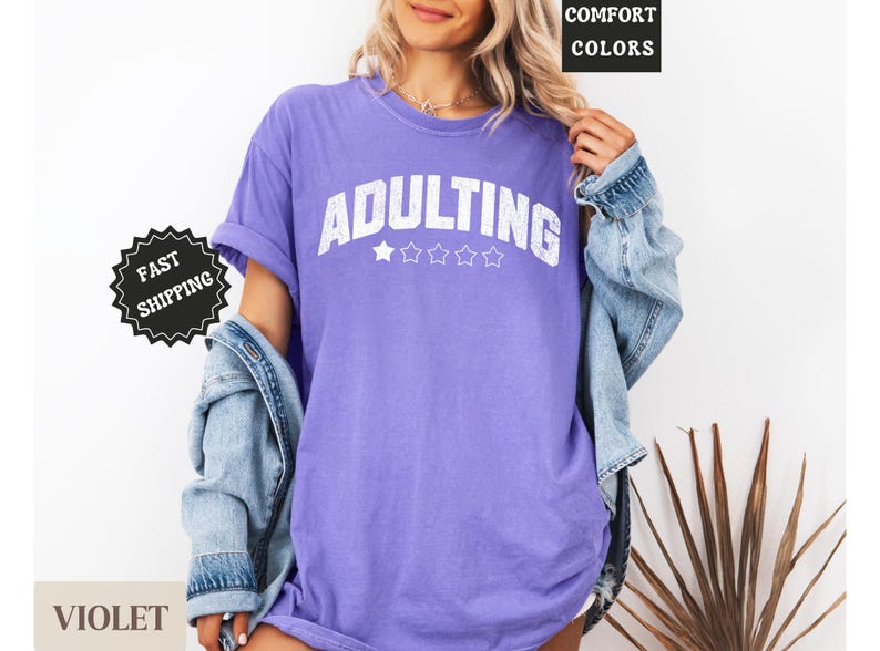 May include: A violet-colored t-shirt with the word "ADULTING" in white block letters. The shirt has five white stars below the word. The model is wearing a denim jacket over the shirt. The words "COMFORT COLORS" are in a black box.