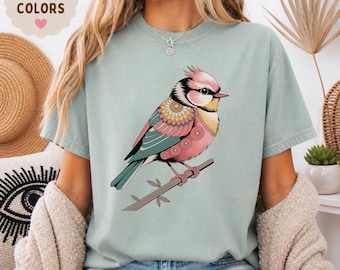 May include: A light green t-shirt featuring a colorful bird illustration. The bird is perched on a branch and has pink, teal, yellow, and black details. The shirt has the words "COMFORT COLORS" and "BAY" printed on it.