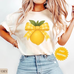 Puede incluir: Una camiseta blanca con un lim&oacute;n amarillo y un lazo de cuadros amarillos y blancos. El texto "ENV&Iacute;O R&Aacute;PIDO" est&aacute; en una pegatina amarilla en forma de estrella.