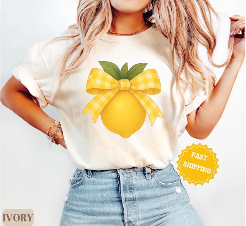 Puede incluir: Una camiseta blanca con un lim&oacute;n amarillo y un lazo de cuadros amarillos y blancos. El texto "ENV&Iacute;O R&Aacute;PIDO" est&aacute; en una pegatina amarilla en forma de estrella.