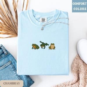 May include: Light blue t-shirt with embroidered frog design. The shirt features three frogs in different poses, in shades of green and brown. A silver necklace with a small pendant rests on the shirt. The words "Comfort Colors" are visible.