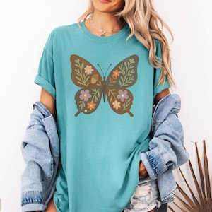 Peut inclure: T-shirt vert d'eau avec un motif de papillon marron orn&eacute; de motifs floraux et de feuilles orange, violet et vert. Le t-shirt a des manches courtes et une coupe d&eacute;contract&eacute;e. Une veste en jean est drap&eacute;e sur les &eacute;paules.