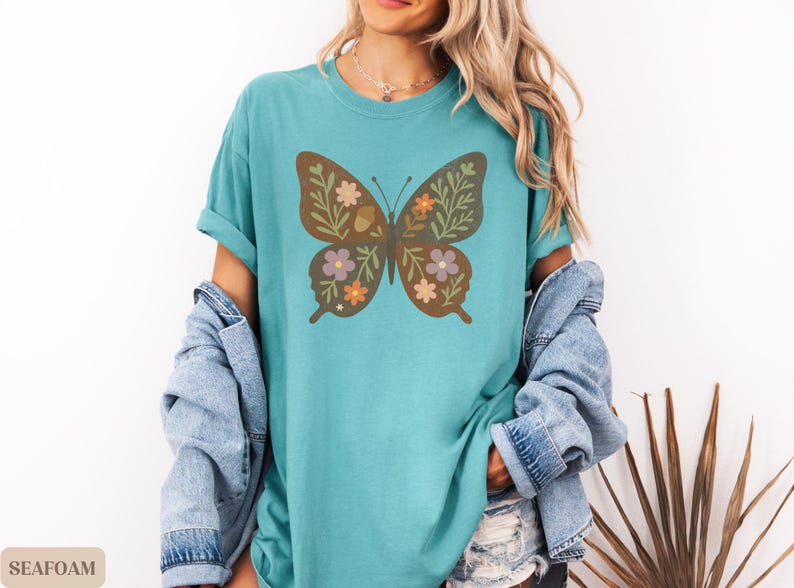Peut inclure: T-shirt vert d'eau avec un motif de papillon marron orn&eacute; de motifs floraux et de feuilles orange, violet et vert. Le t-shirt a des manches courtes et une coupe d&eacute;contract&eacute;e. Une veste en jean est drap&eacute;e sur les &eacute;paules.