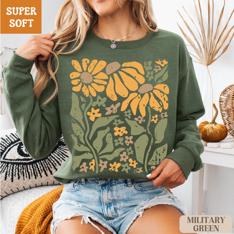May include: Olive green sweatshirt with a floral design featuring large yellow and brown sunflowers and smaller flowers. The words "SUPER SOFT" are in a brown box in the upper left corner. The words "MILITARY GREEN" are in a brown box in the lower right corner.