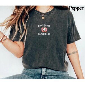 May include: A dark grey t-shirt with a white graphic of a goose sitting in a pink armchair. The text "Silly Goose Psych Club" is printed below the graphic.