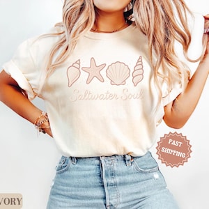 May include: Ivory-colored t-shirt featuring a seashell design and the words "Saltwater Soul" in a script font. The design includes a starfish and various seashell illustrations. The shirt is paired with blue jeans.