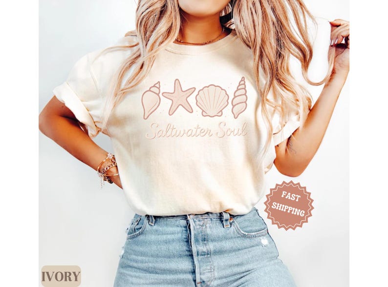 May include: Ivory-colored t-shirt featuring a seashell design and the words "Saltwater Soul" in a script font. The design includes a starfish and various seashell illustrations. The shirt is paired with blue jeans.