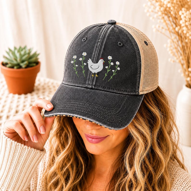 May include: A black and tan trucker hat with a chicken and floral embroidery. The front panel is black with a distressed look, and the mesh back is tan. The chicken is white with yellow feet and a red comb, surrounded by white flowers.