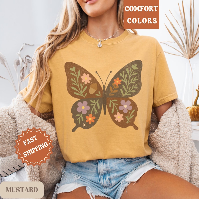 Peut inclure: T-shirt moutarde avec un grand motif de papillon rempli de fleurs et de feuilles. Le papillon est marron avec des accents orange, roses et violets. Le t-shirt porte &eacute;galement les mots "Comfort Colors" et "Fast Shipping".