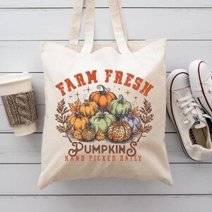 May include: A white canvas tote bag with the text "Farm Fresh Pumpkins Hand Picked Daily" in orange and brown. The bag features a vintage illustration of pumpkins in various colors and sizes.