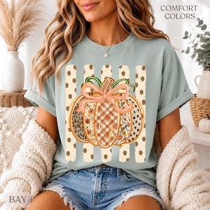 May include: A light sage green short-sleeve t-shirt featuring a central graphic of a pumpkin with a pink bow. The pumpkin has segments with leopard print, plaid, and floral patterns. Behind the pumpkin are vertical beige stripes with brown dots.
