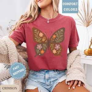 Peut inclure: T-shirt rouge cramoisi avec un motif de papillon orn&eacute; de motifs floraux et de glands. Le t-shirt fait partie de la collection "Comfort Colors" et est associ&eacute; &agrave; un short en jean. Un pull cr&egrave;me est &eacute;galement visible.