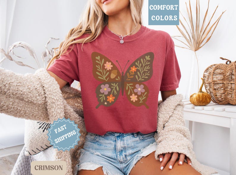 Peut inclure: T-shirt rouge cramoisi avec un motif de papillon orn&eacute; de motifs floraux et de glands. Le t-shirt fait partie de la collection "Comfort Colors" et est associ&eacute; &agrave; un short en jean. Un pull cr&egrave;me est &eacute;galement visible.