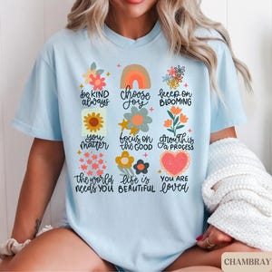 Pu&ograve; includere: Maglietta blu chiaro con un design floreale e affermazioni positive. Il testo recita: "Sii gentile sempre, scegli la gioia, continua a fiorire, tu conti, concentrati sul bene, la crescita &egrave; un processo, il mondo ha bisogno di te, la vita &egrave; bella, sei amato."