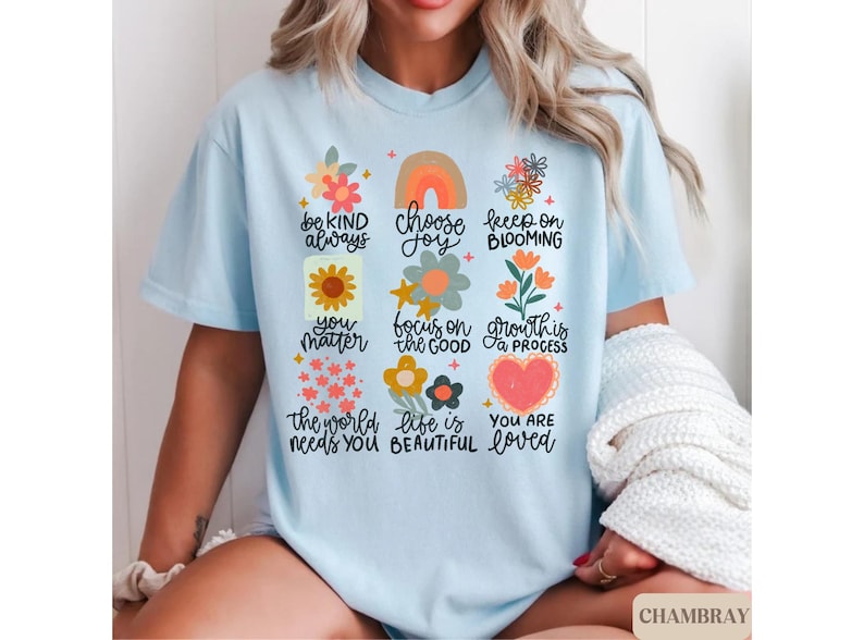 Pu&ograve; includere: Maglietta blu chiaro con un design floreale e affermazioni positive. Il testo recita: "Sii gentile sempre, scegli la gioia, continua a fiorire, tu conti, concentrati sul bene, la crescita &egrave; un processo, il mondo ha bisogno di te, la vita &egrave; bella, sei amato."