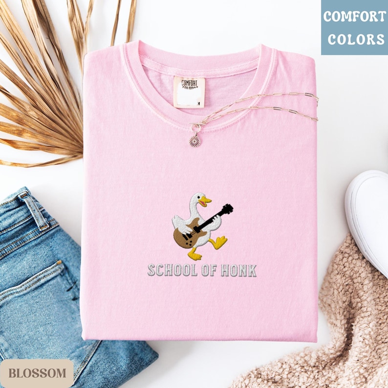 May include: A light pink t-shirt with a round neck, featuring an embroidered cartoon goose playing a guitar. The text "SCHOOL OF HONK" is below the goose. A delicate necklace with a pendant rests on the shirt. The shirt is folded on a white surface.