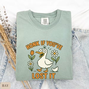 May include: A light green Comfort Colors t-shirt with the phrase "HONK IF YOU'VE LOST IT" in orange lettering. The shirt features a cartoon duck with wide eyes, surrounded by flowers. The shirt is folded on top of a pair of light blue jeans.