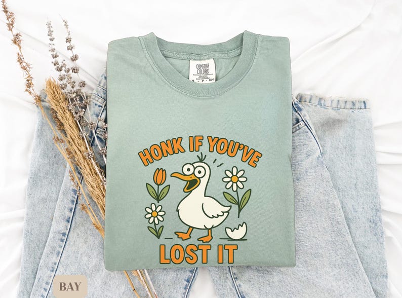 May include: A light green Comfort Colors t-shirt with the phrase "HONK IF YOU'VE LOST IT" in orange lettering. The shirt features a cartoon duck with wide eyes, surrounded by flowers. The shirt is folded on top of a pair of light blue jeans.