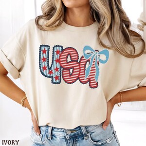 Op de afbeelding: Ivoor kleurig t-shirt met het woord "USA" in een patriottisch ontwerp. De letters zijn rood, wit en blauw, met sterren en strepen. Een lichtblauwe strik staat op de letter "A".