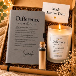 May include: A gift box containing a gray notebook with the text Difference maker and the name Sarah, a lit candle in a frosted glass jar with a label reading Smells Like Difference maker, and a small glass bottle filled with wooden matches.