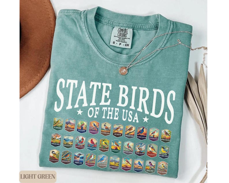 May include: Light green t-shirt with the text "State Birds of the USA" in white. The shirt features a graphic of a collection of colorful state birds in vintage-style illustrations.