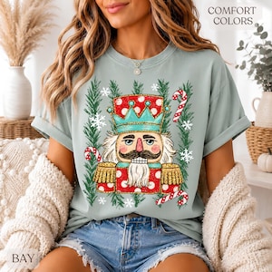 Könnte beinhalten: Ein helles salbeigrünes T-Shirt mit einer Grafik eines Nussknackers in einer rot-türkisfarbenen Uniform mit goldenen Schulterklappen und Schnurrbart. Das Motiv ist von grünen Tannenzweigen, weißen Schneeflocken und Zuckerstangen umgeben.