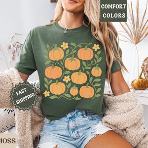 May include: Green t-shirt featuring a pumpkin design. The shirt has a crew neck and short sleeves. The pumpkin graphic is orange with green leaves and yellow flowers. The shirt is made of a soft, comfortable material.
