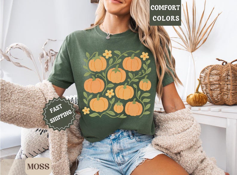 May include: Green t-shirt featuring a pumpkin design. The shirt has a crew neck and short sleeves. The pumpkin graphic is orange with green leaves and yellow flowers. The shirt is made of a soft, comfortable material.