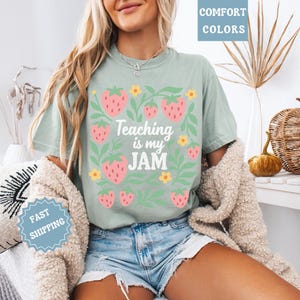 May include: Sage green t-shirt with a strawberry and floral design. The text "Teaching is my JAM" is printed in white. The shirt is part of the comfort colors collection. The model is wearing denim shorts and a beige cardigan.