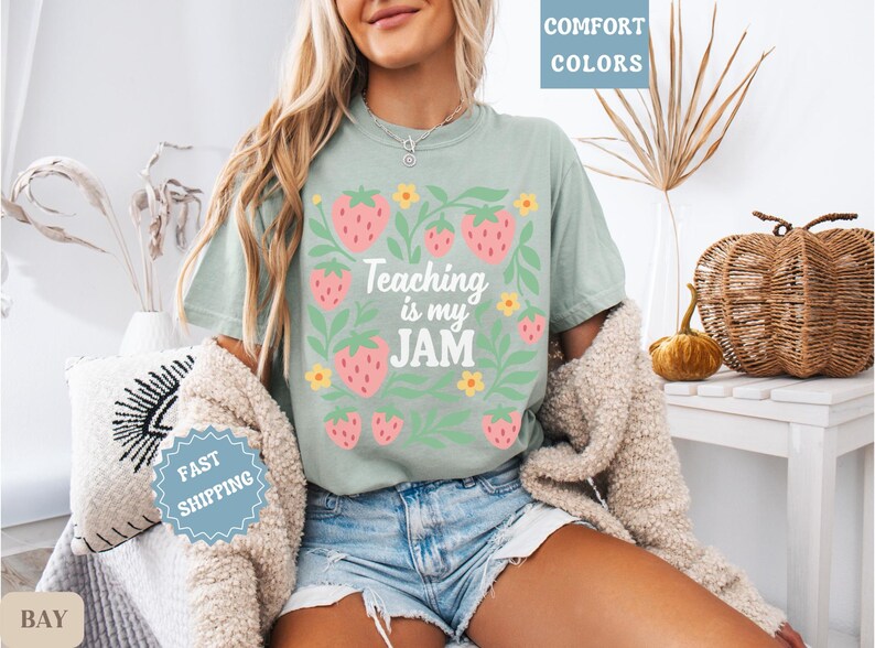 May include: Sage green t-shirt with a strawberry and floral design. The text "Teaching is my JAM" is printed in white. The shirt is part of the comfort colors collection. The model is wearing denim shorts and a beige cardigan.